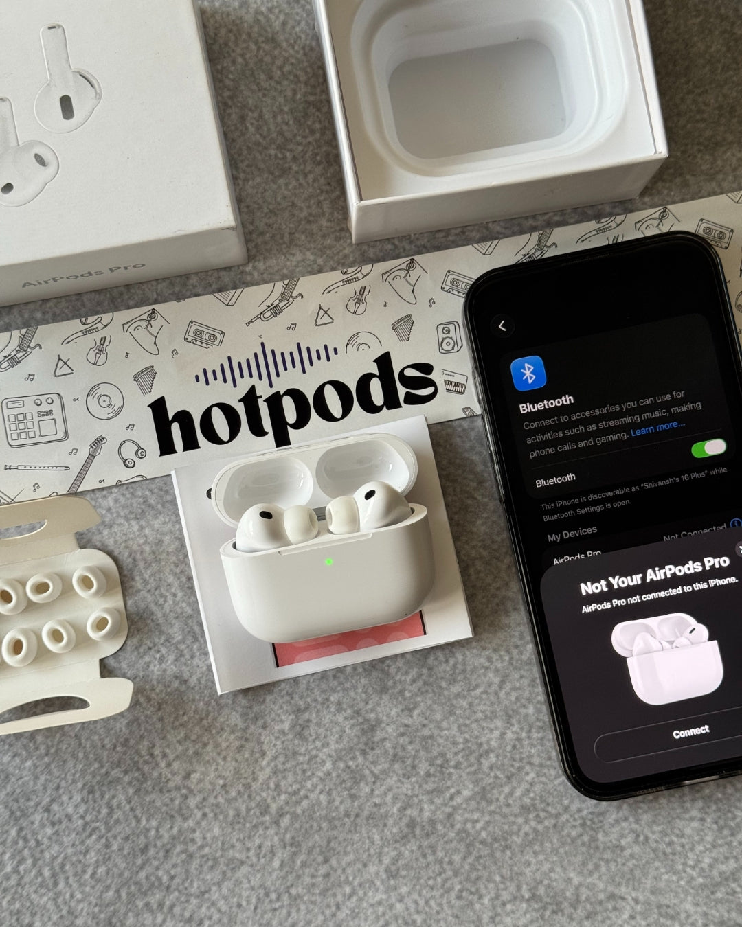 AirPods Pro with charging case, phone showing Bluetooth connection screen on a branded 'hotpods' mat.
